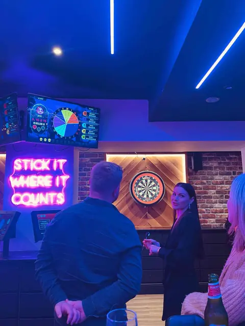 A darts night under neon lights, a scoreboard screen above the oche.