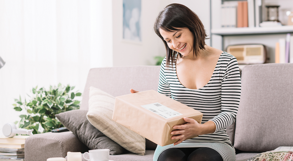 Woman sitting on a sofa at home, holding a recently delivered parcel and looking down at the package.